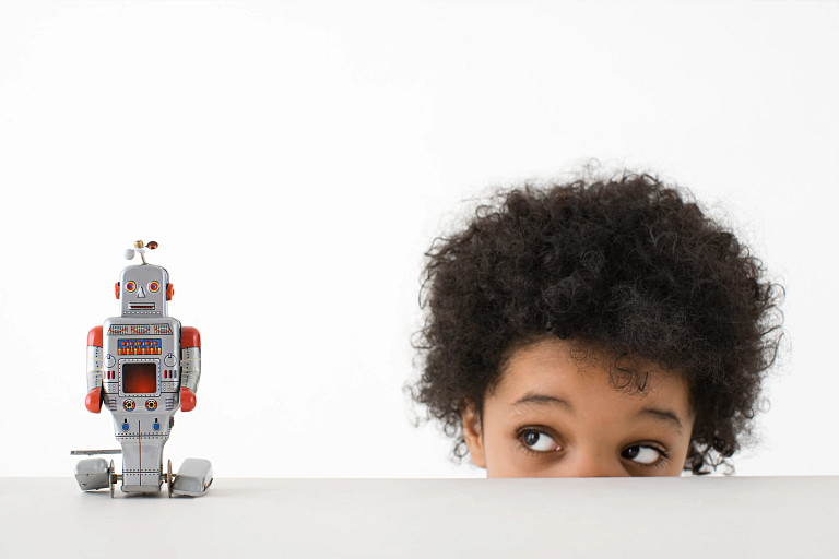 Vintage silver toy robot with red accents standing on a table, being observed by a child with curly black hair standing behind the table.